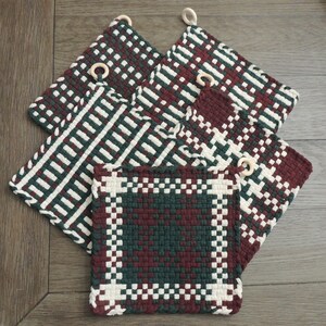 Peut inclure: Quatre maniques tissées dans un motif vert, blanc et bordeaux. Chaque manique est dotée d'une bague en bois fixée à un coin.