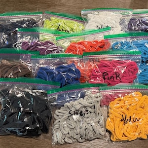 May include: A collection of clear plastic zip-top bags filled with various colors of coiled fabric loops. The bags contain loops in black, brown, purple, lime green, white, red, blue, pink, silver, and yellow, with some bags labeled in black marker.