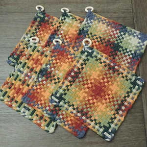 May include: A stack of five woven potholders. Each potholder is square with a colorful plaid pattern in shades of orange, red, green, blue, and yellow. Each potholder has a white loop for hanging.