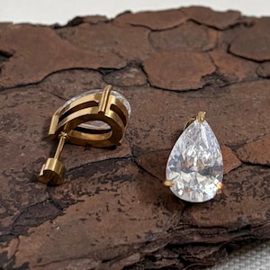 May include: A pair of gold-tone stud earrings featuring a clear, pear-shaped faceted gemstone. One earring is positioned upright to show the teardrop-shaped stone, while the other is placed on its side to reveal the gold-tone post and backing.
