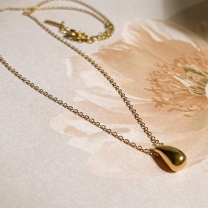 May include: A gold-colored necklace with a teardrop pendant. The chain is delicate and the pendant is smooth and rounded. The necklace is displayed on a light-colored surface with a floral design in the background.