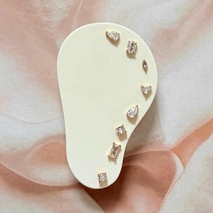 May include: A set of eight gold-toned stud earrings featuring clear, faceted stones in various shapes including pear, marquise, round, and rectangular cuts, all arranged in a curved line on a cream-coloured, ear-shaped display card.