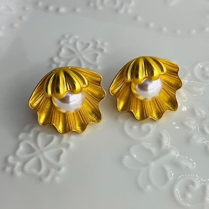 May include: A pair of gold-coloured seashell stud earrings, each with a white pearl centre. The earrings have a detailed, textured design, set against a white background.