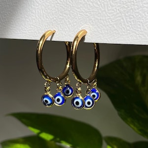 May include: Gold hoop earrings with multiple dangling blue evil eye charms. The earrings are a popular style of jewelry.
