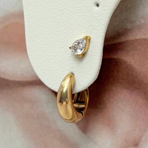 Peut inclure: Un ensemble de boucles d'oreilles dorées composé d'une petite créole épaisse et lisse, ainsi qu'une puce d'oreille ornée d'une pierre transparente en forme de poire sertie dans une monture dorée, le tout présenté sur un support blanc.