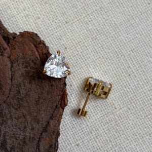May include: A pair of stud earrings featuring clear, heart-shaped faceted stones set in gold-toned metal. One earring is positioned on a piece of dark brown bark, while the other is placed nearby, showing the post and backing mechanism clearly.