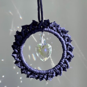 May include: A navy blue macrame wreath with a heart-shaped crystal pendant. The wreath is circular, with a textured, knotted design. The crystal heart hangs in the center, reflecting light. A navy blue cord is attached for hanging.