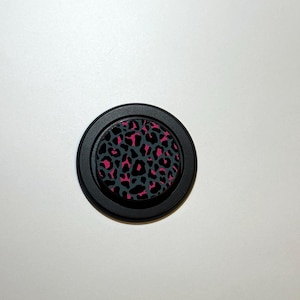 May include: A black circular phone grip with a pink and gray leopard print design. The grip has a raised, textured surface and a black border. The phone accessory is set against a white background.