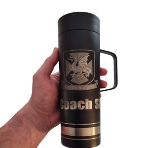 May include: Black insulated travel mug with a handle, featuring a silver emblem of a dragon and the text "Clevedon Utd JFC" and "Coach Si". The mug has a silver band near the bottom.