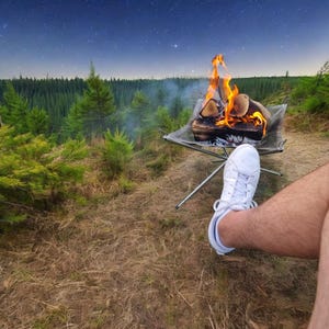 May include: A campfire with burning logs in a portable fire pit. The flames are bright orange and yellow. A person's white trainer is in the foreground. The background features a forest and a starry night sky.