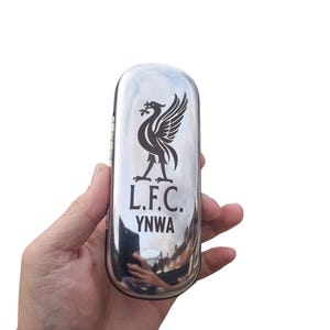 Może przedstawiać: Polerowane, srebrne etui z emblematem Liverpool Football Club. Emblemat zawiera ptaka Liver oraz czarne litery „L.F.C.” i „YNWA”. Etui ma odblaskową powierzchnię.