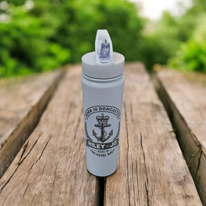 May include: A light gray water bottle with a white flip-top lid. The bottle features a black graphic with an anchor, crown, and text that reads "BORN IN DONCASTER" and "RILEY-JO MADE IN THE ROYAL NAVY".