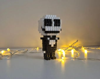 May include: A small, three-dimensional character figurine constructed from black and white square beads. The figure features a large, rounded head with black bead eyes and a wide, stitched-style mouth, set atop a slender black body with a white collar.