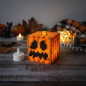 May include: A glowing orange and black Halloween pumpkin decoration made of small beads. The pumpkin has a jack-o'-lantern face and a yellow stem. Other Halloween decorations, including candles and a black cat figurine, are in the background.