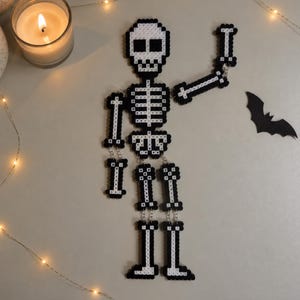Könnte beinhalten: Eine schwarz-weiße Skelettfigur aus kleinen quadratischen Perlen. Das Skelett besteht aus einem Schädel, einem Brustkorb, einem Becken und beweglichen Gliedmaßen, die mit silbernen Metallringen verbunden sind. Ein Arm ist erhoben.
