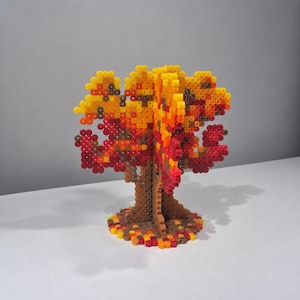 May include: A three-dimensional tree sculpture constructed from interlocking plastic fuse beads. The tree features a brown trunk and a canopy composed of yellow, orange, and red beads, with a circular base covered in scattered fallen leaf-coloured beads.
