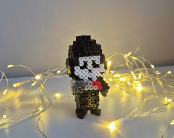 May include: A three-dimensional pixel art figurine constructed from black, white, gray, and red plastic fuse beads. The figure features a black head and body, a white mask-like face, and small red and gray accents on the front of the torso.