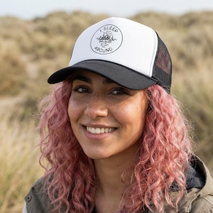 Puede incluir: Gorra de camionero en blanco y negro con el texto "I SLEEP AROUND" en un diseño circular. La gorra tiene un panel frontal blanco, una visera negra y una parte trasera de malla negra. El diseño incluye una ilustración de montaña y árboles.
