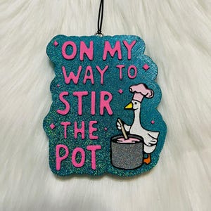 May include: A teal blue glitter ornament with a pink and white cartoon goose stirring a pot with a wooden spoon. The ornament has the text "On my way to stir the pot" in pink lettering.