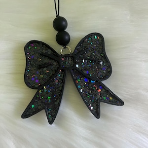 May include: Black glitter bow-shaped car charm with iridescent accents. The bow is suspended from a black cord with two black spherical beads. The charm is set against a white, fluffy background.