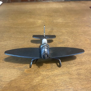 以下が含まれることがあります： スパークプラグと金属板で作られた小さな金属製の飛行機の彫刻。飛行機は黒で、スパークプラグは白です。