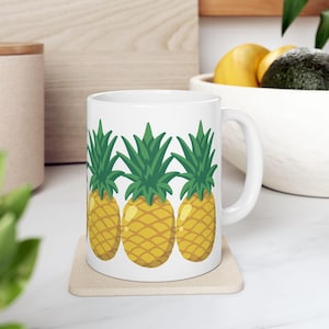 Peut inclure: Tasse en céramique blanche avec un motif répété d'ananas jaunes et verts. La tasse a une anse incurvée et repose sur un dessous de verre beige. L'arrière-plan comprend une planche à découper en bois et un bol de fruits.