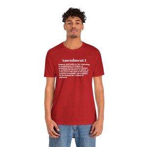 Puede incluir: Una camiseta roja con el texto "Amendment I" y el texto de la Primera Enmienda de la Constitución de los Estados Unidos en blanco. La camiseta es de cuello redondo.