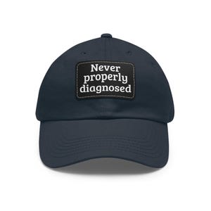 Può includere: Un cappellino da baseball blu navy con una toppa rettangolare nera sul davanti. La toppa mostra il testo bianco "Never properly diagnosed" in un carattere serif. Il cappellino presenta una visiera curva e cuciture sulla corona e visiera.