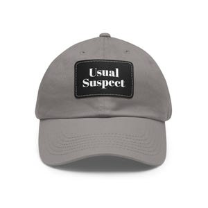 May include: A grey baseball cap featuring a rectangular black patch centered on the front. The patch displays the white text "Usual Suspect" in a serif font. The cap has a curved brim, a button on the top, and visible stitching along the panels.