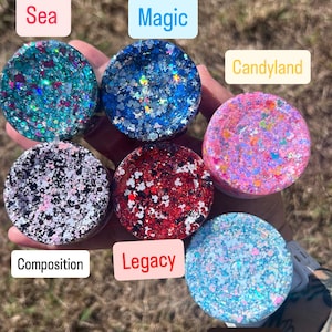 May include: A collection of six small, round containers filled with colorful glitter. Each container is labeled with a different word: Sea, Magic, Candyland, Composition, Legacy, and Sleepy. The glitter colors vary, including blue, red, pink, and black.