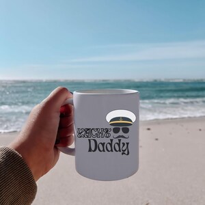 May include: White ceramic mug with a black and white illustration of a captain's hat, sunglasses, and mustache. The text "Yacht Daddy" is printed in black below the illustration.