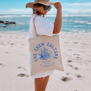 Op de afbeelding: Een beige tote bag met een blauwe afbeelding van een schip en de tekst "THE CREW CULTURE YACHT DADDY FORT LAUDERDALE, USA".