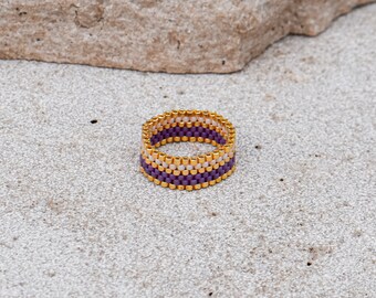 Op de afbeelding: Een ring van paarse, witte en gouden kralen. De ring is gemaakt van kleine, vierkante kralen die in een patroon aan elkaar zijn geweven.