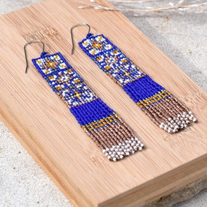 Op de afbeelding: Een paar lange, geperde oorbellen met een geometrisch patroon in tinten blauw, wit, goud en bruin. De oorbellen hangen aan zilveren haakjes.