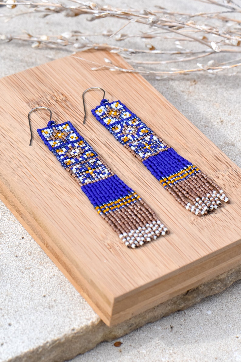 Op de afbeelding: Een paar lange, geperde oorbellen met een geometrisch patroon in tinten blauw, wit, goud en bruin. De oorbellen hangen aan zilveren haakjes.