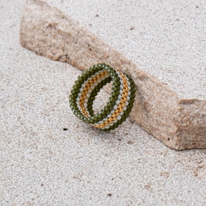 May include: A green, white, and gold beaded ring. The ring is made of small beads that are woven together to create a striped pattern.