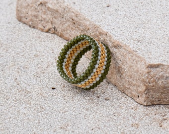 Op de afbeelding: Een groene, witte en gouden kralenring. De ring is gemaakt van kleine kralen die samen geweven zijn om een gestreept patroon te creëren.