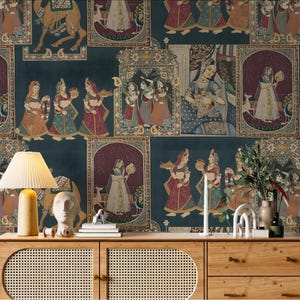 May include: Wallpaper featuring a repeating pattern of framed illustrations depicting figures in traditional Indian attire, camels, and deer against a dark teal background. The scene is set on a wooden cabinet with a lamp and decorative items.