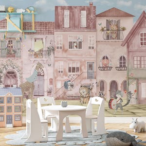 A custom wallpaper for walls displayed as a nursery wall mural in a bright playroom with a white craft table and chairs. The whimsical animal village design fills the entire back wall, creating a storybook atmosphere. The scale shows how the watercolor townhouses and musical animals interact with kid-sized furniture.