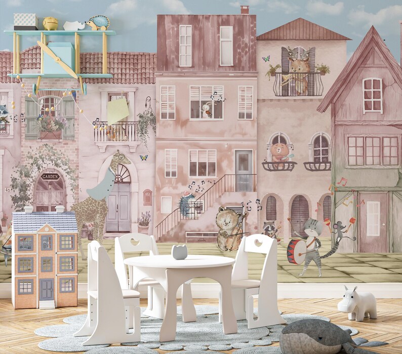 A custom wallpaper for walls displayed as a nursery wall mural in a bright playroom with a white craft table and chairs. The whimsical animal village design fills the entire back wall, creating a storybook atmosphere. The scale shows how the watercolor townhouses and musical animals interact with kid-sized furniture.