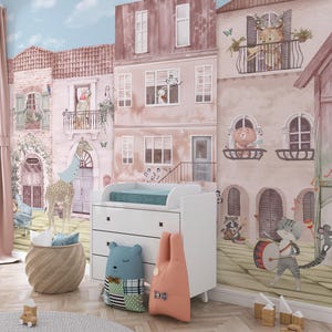 An interior mockup of a custom wallpaper for walls used in a modern nursery next to a white changing table and a blue bear-shaped pillow. The angle highlights the watercolor detail of the townhouses and the musical cat drummer, demonstrating a high-end, designer finish for a gender-neutral baby room.