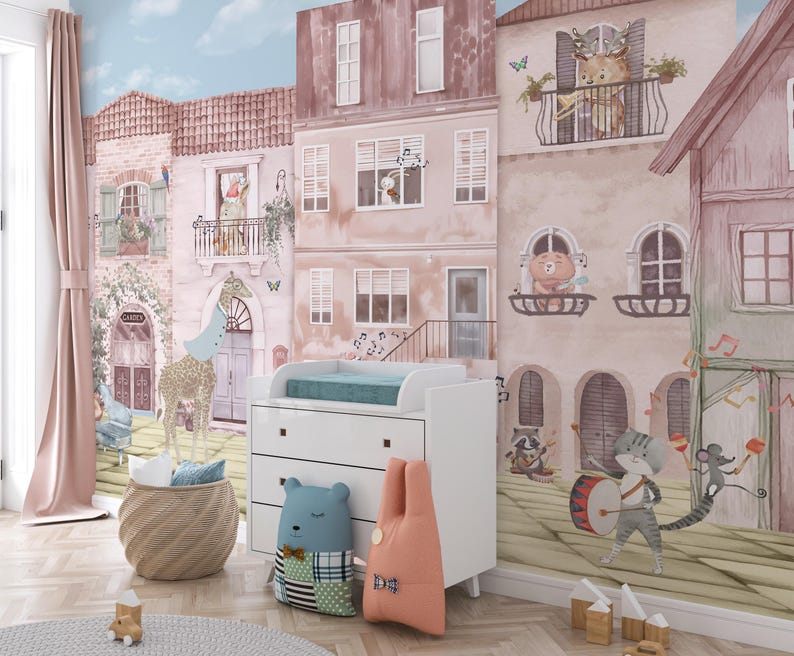 An interior mockup of a custom wallpaper for walls used in a modern nursery next to a white changing table and a blue bear-shaped pillow. The angle highlights the watercolor detail of the townhouses and the musical cat drummer, demonstrating a high-end, designer finish for a gender-neutral baby room.