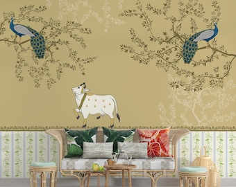 May include: A white cow with black spots stands in front of a couch with patterned pillows. The wall behind the couch features a gold background with a repeating pattern of stylized trees and peacocks.
