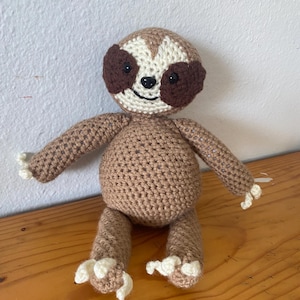 May include: A crocheted brown sloth toy with white claws and a white face with brown eyes.