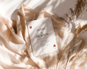 Könnte beinhalten: Eine weiße Hochzeitseinladung mit schwarzem Text, der "Avery & Jackson" lautet, und einem roten Herzsymbol. Die Einladung liegt auf einem beigefarbenen Stoffhintergrund mit getrocknetem Gras.