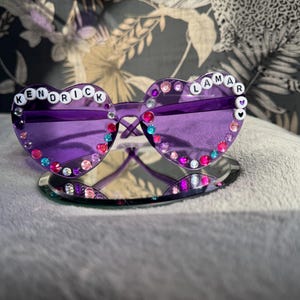 Puede incluir: Gafas de sol de corazón morado con pedrería y las palabras "Kendrick Lamar" escritas en letras blancas.