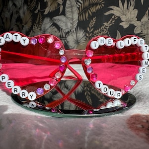 May include: Pink heart-shaped sunglasses with white beads spelling "Katy Perry The Life Times Tour"