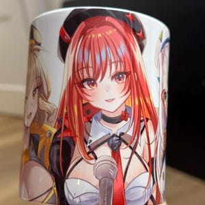 Puede incluir: Taza de cerámica blanca con ilustraciones de estilo anime de tres personajes femeninos. El personaje central tiene el pelo largo rojo, una boina negra y una corbata roja. Un micrófono está frente al personaje. Los otros personajes tienen el pelo rubio y blanco.