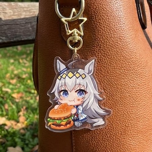May include: A clear acrylic keyring featuring a cartoon character with white hair and blue eyes, eating a burger. The keyring is attached to a gold-coloured star-shaped clasp and a brown leather bag.