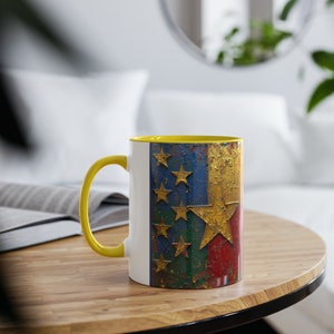 May include: A white ceramic mug with a yellow rim and handle. The mug features a distressed, rusty design of the flag of Kosovo. The flag is blue, red, and green with six yellow stars and a large yellow star in the centre.